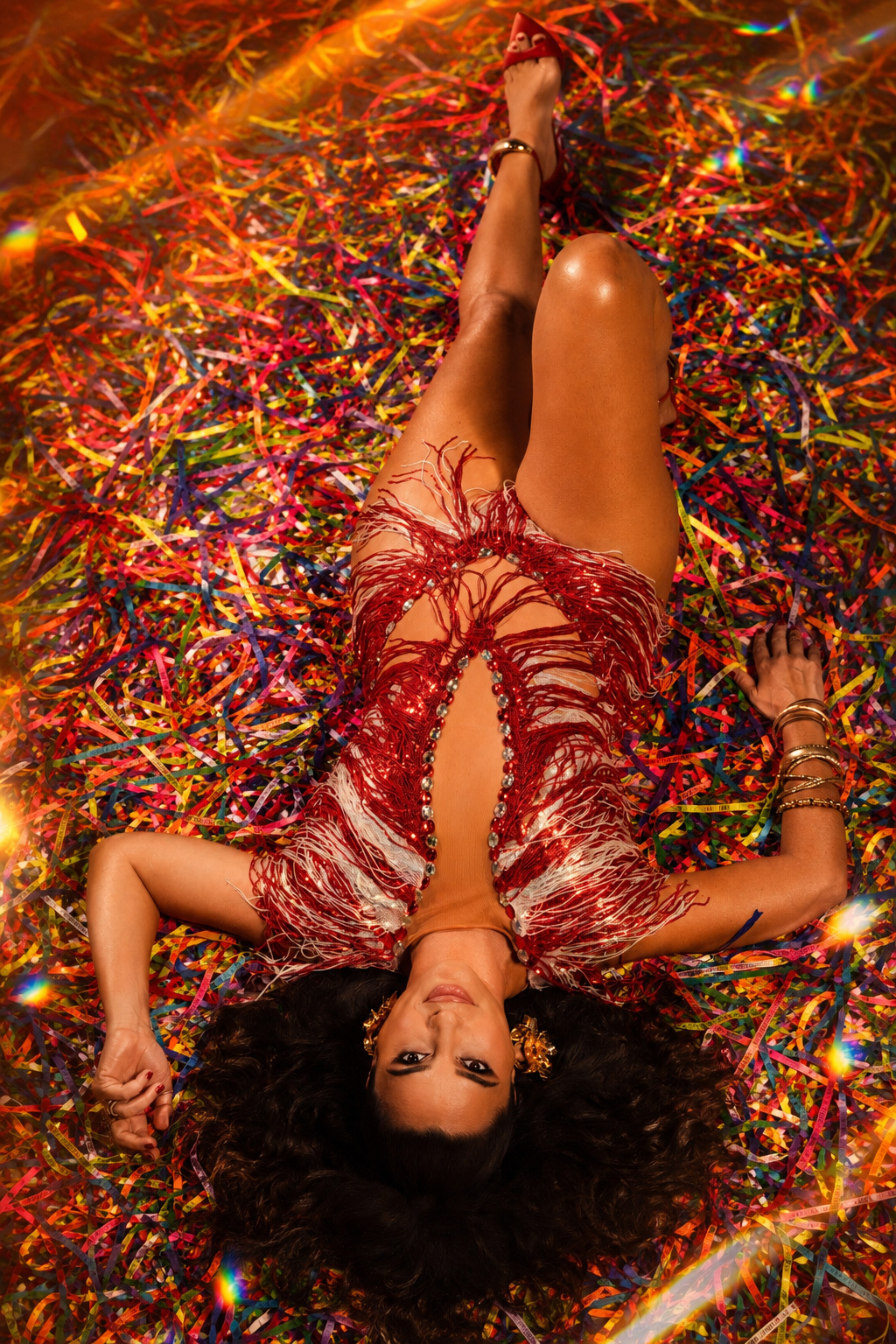 Aline Calixto deitada sobre um tapete denso de fitas de carnaval coloridas, vista de cima, vestido com franjas vermelhas e brancas, expressão serena, luz quente em volta.