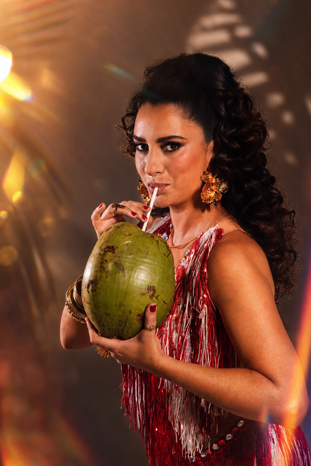 Aline Calixto em pé com um coco verde nas mãos, perna direita levantada, vestido com franjas vermelhas, sombras de palmeira ao fundo, atmosfera solar em tom bege.