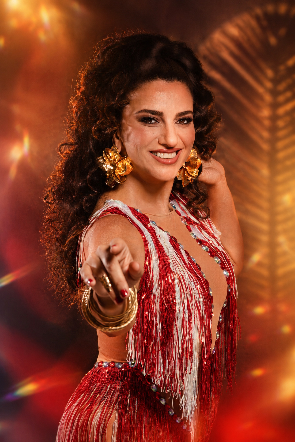 Close de Aline Calixto sorrindo, mão estendida à frente com braceletes dourados, brincos de flor dourados, vestido com franjas vermelhas e brancas, fundo dourado com sombra de palmeira.