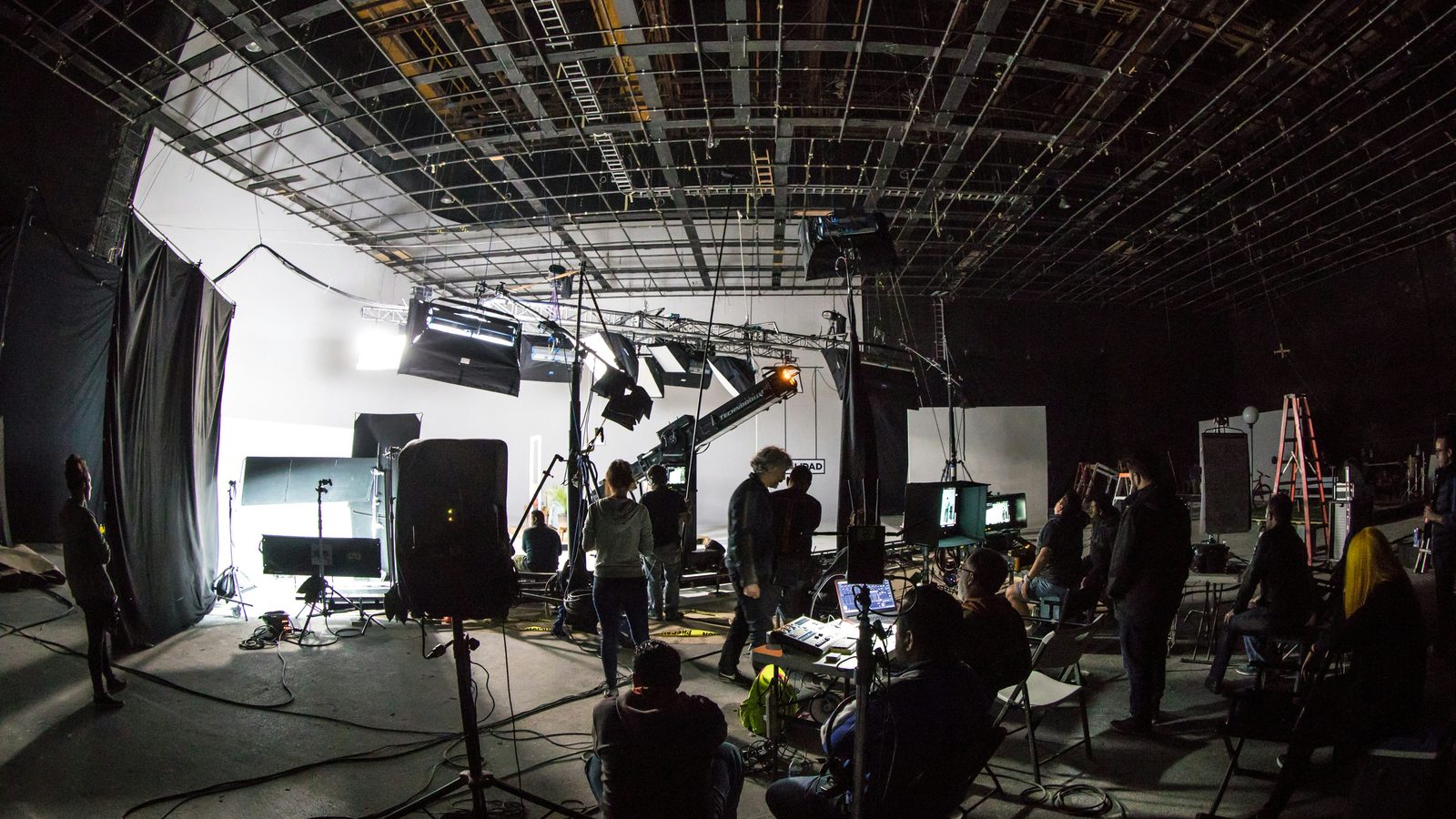 Set de produção audiovisual profissional em um galpão amplo, com equipe técnica, refletores em treliça, monitores e cabos.