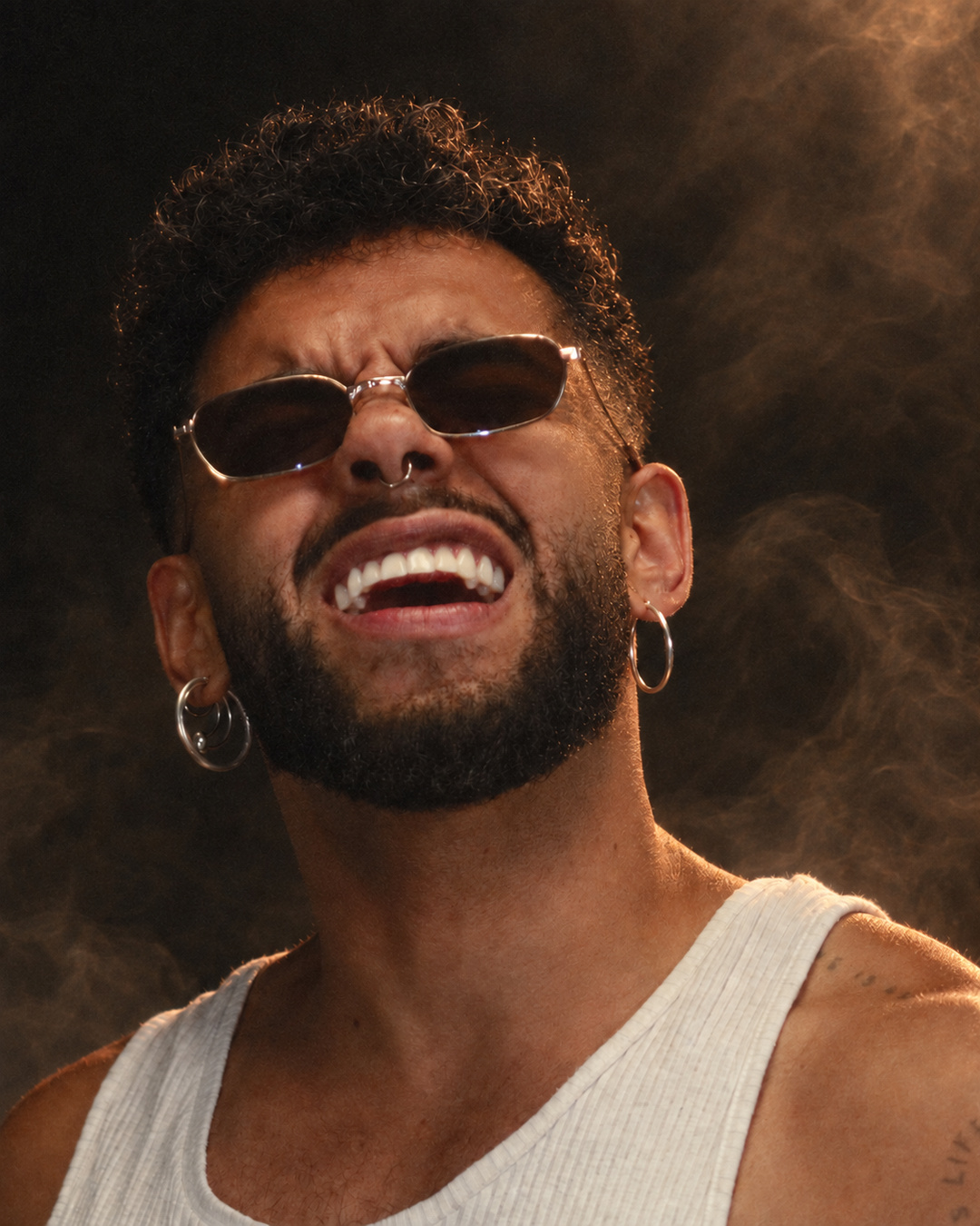Close de Saulo sorrindo, com óculos escuros e luz quente nos cabelos, diante de fundo com fumaça.